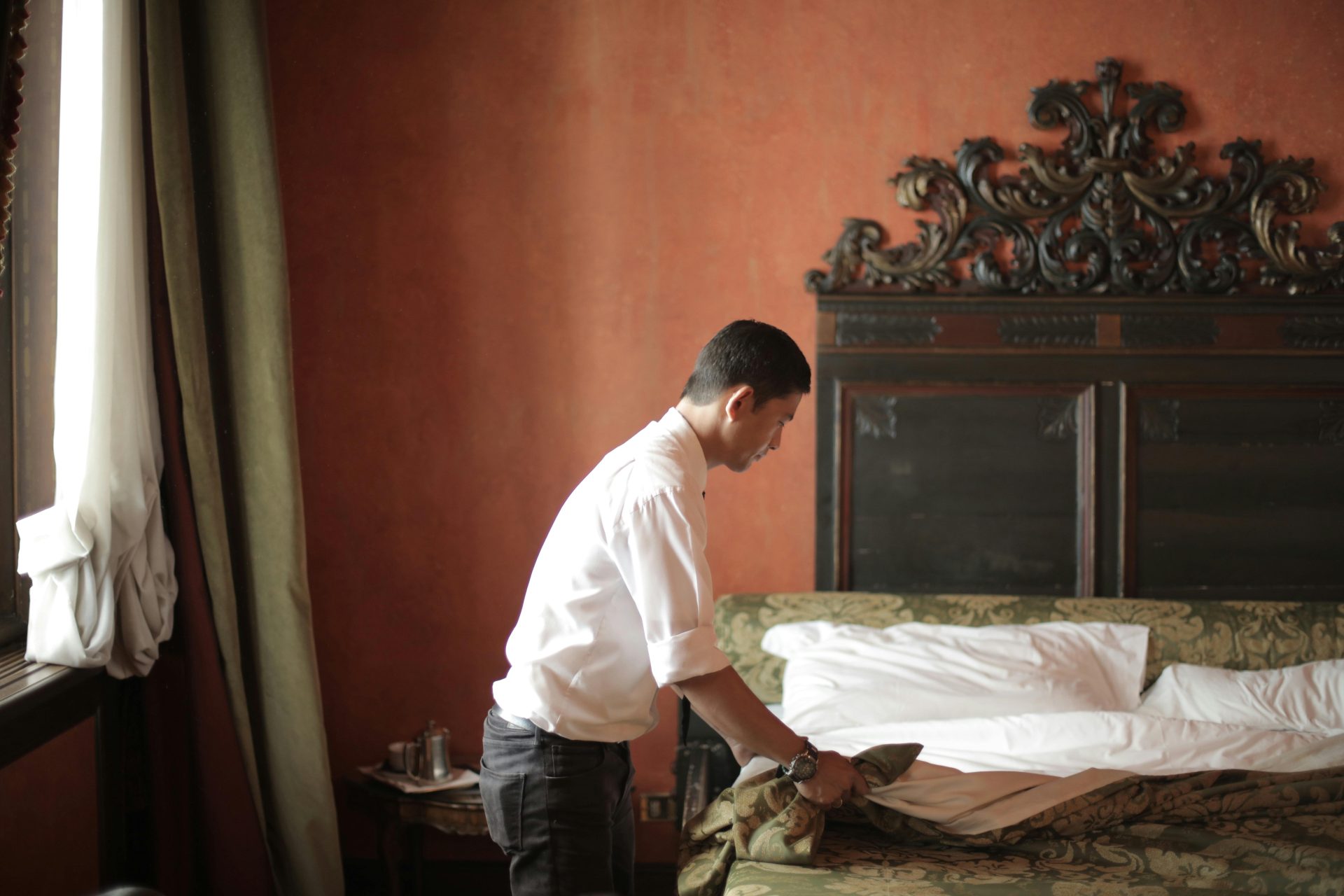 Side view of young male housekeeper making bed while arranging stylish hotel bedroom with vintage interior